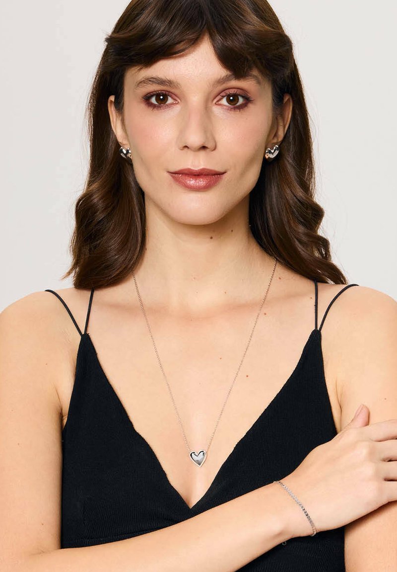 Srebrny zestaw biżuterii, w tym naszyjnik z serduszkowym wisiorkiem oraz pasujące kolczyki. Modelka ma na sobie czarny top na ramiączkach.