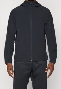 Homme portant une veste à capuche noire zippée intégralement et un pantalon de survêtement noir, tous deux avec un petit logo Nike sur la poitrine et la cuisse.