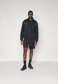 Veste de sport noire avec des rayures vertes, fermeture éclair, et shorts en maille. Accents de logo en vert. Tenue complétée par des chaussures et des chaussettes noires.