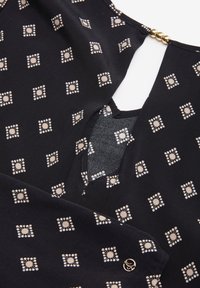 Svart tyg med beige geometriskt prickmönster, med en triangulär halsringning med gulddetaljer och ett litet runt guldemblem på ärmen.