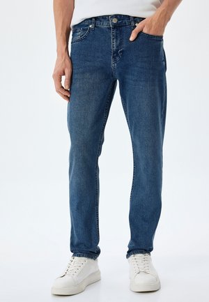 Dunkelblaue Jeans aus Denim, mit geradem Schnitt, fünf Taschen-Design und subtilen Abnutzungen auf dem Stoff. Getragen mit weißen Sneakern.