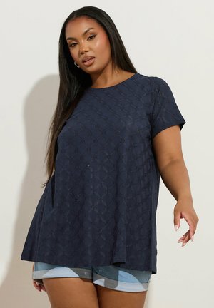 Kobieta z długimi ciemnymi włosami, ubrana w granatową bluzkę w wzory z krótkim rękawem oraz jasne krótkie spodenki jeansowe, na jednolitym tle.