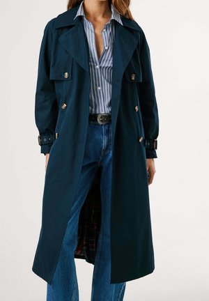 Kvinde iført en lang mørk trenchcoat over en blå- og hvidstribet skjorte, kombineret med blå jeans og et sort bælte med en dekorativ spænde.