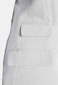 Tecido cinza claro com um acabamento texturizado. Apresenta dois bolsos retangulares, planos, cosidos na costura lateral.