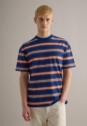 Jonge man met blond haar, gekleed in een blauw, rood en wit gestreept T-shirt met korte mouwen en een voorzak, en lichte broek.