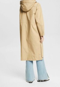 Persona che indossa un lungo cappotto beige con cappuccio e fessure laterali, insieme a jeans sfilacciati di un blu chiaro, in piedi su uno sfondo neutro.