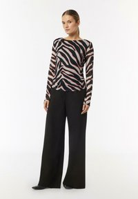 Langärmliges Oberteil aus durchsichtigen Stoff mit einem schwarz-pinken Zebra-Streifenmuster, kombiniert mit weiten schwarzen Hosen und schwarzen Schuhen.