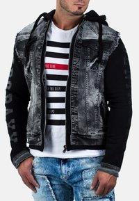 Giacca in denim con maniche nere, corpo grigio lavato, chiusura con zip e cappuccio. Presenta dettagli strappati e un logo sul braccio.