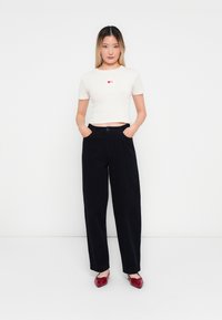 Witte geribbelde crop top met rode hartafbeelding, gecombineerd met zwarte wijde broek en glanzende rode loafers. Eenvoudige, casual look.