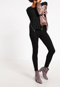 Svart stickad tröja, taupe bomberjacka, smala svarta byxor, grå ankelstövlar med dragkedjor, på en vit bakgrund.