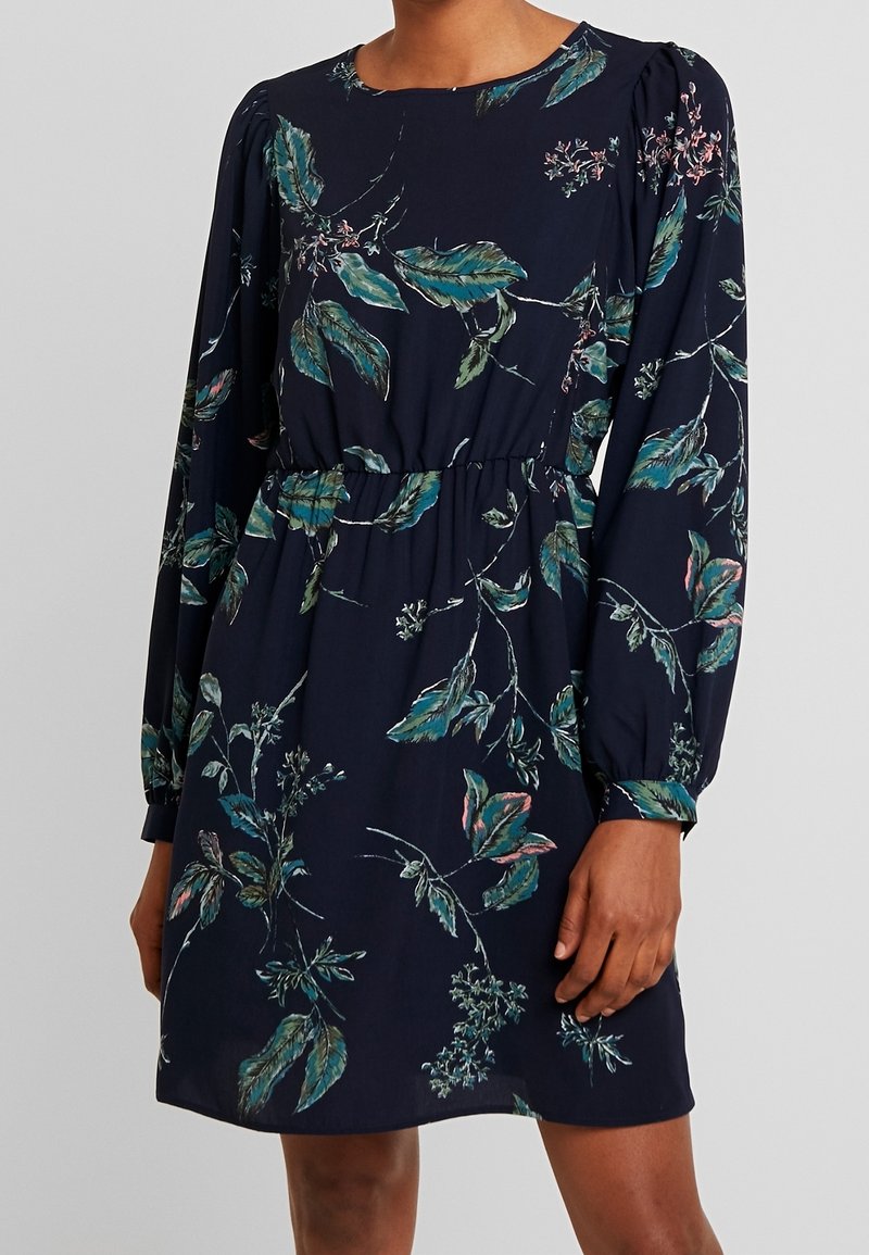 Květinové šaty v tmavě námořnické barvě s dlouhými rukávy, zdobené zelenými a růžovými botanickými vzory, s úzkým pasem a kulatým výstřihem.
