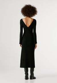 Juoda nertinė suknelė su giliu V formos nugara, ilgos rankovės ir prigludusi siluetas. Lygus audinys ir iki kulkšnies siekianti apatinė dalis. Dėvima su juodomis ilgoji batais iki kelių.