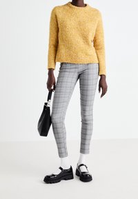 Persoon in een gele textuurtrui, grijze geplooide slim fit broek, witte sokken, zwarte chunky Mary Jane schoenen, met een zwarte handtas.