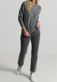 Femme portant un pull gris à torsades, un jean droit gris foncé, des baskets blanches et des bracelets, marchant devant un fond uni.