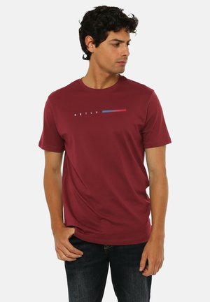 Uomo con capelli ricci scuri che indossa una camicia bordeaux a maniche corte con righe orizzontali blu e rosse e pantaloni neri, in piedi davanti a uno sfondo bianco.