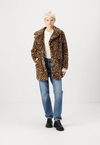Leopardenprint nepbontjas met een brede kraag, knoopsluiting aan de voorkant en zijzakken, gedragen over een lichte gebreide trui en blauwe spijkerbroek.