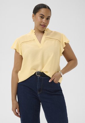 Vrouw met een lichtgele blouse met gerimpelde mouwen en kanten details, donkerblauwe spijkerbroek, een zwarte riem en een zilveren armband, poserend met één hand in de zak.