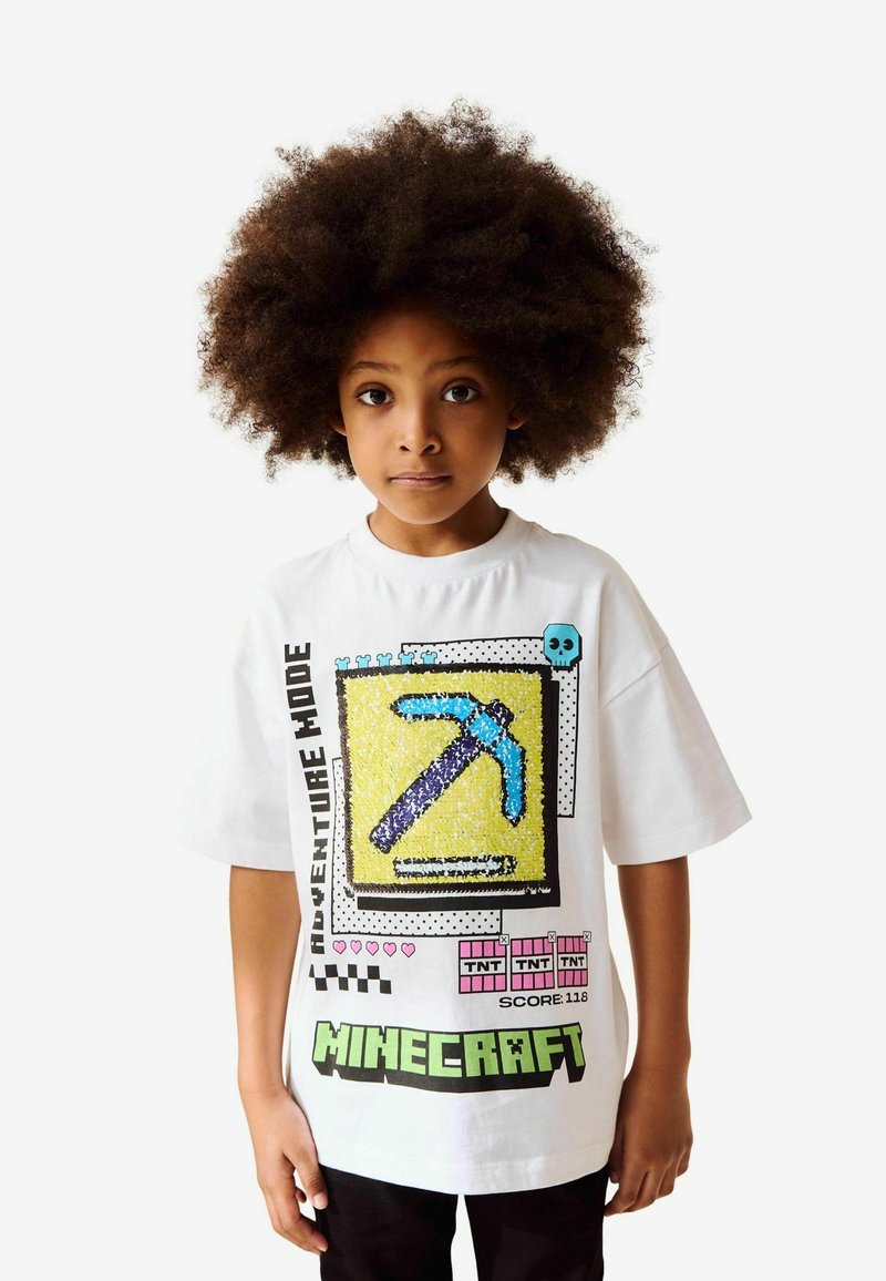T-shirt bianco con un design in pixel art su sfondo giallo, piccone blu, testo nero e grafiche colorate ispirate ai giochi.