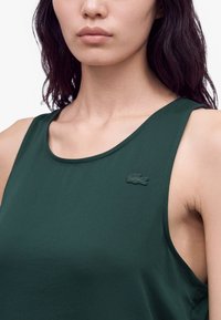 Femme portant un haut sans manches vert foncé avec un petit logo crocodile brodé sur le côté gauche près de l'épaule.