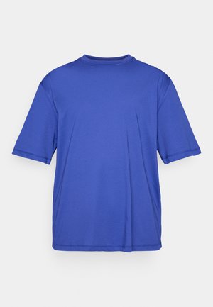 T-shirt azul de manga curta, com um corte solto e um discreto detalhe de logótipo no peito, contra um fundo claro e simples.