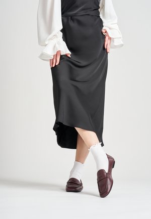Jupe en satin noir avec un ourlet en forme de scoop, associée à un chemisier blanc et des mocassins en cuir bordeaux sur des chaussettes blanches à volants.