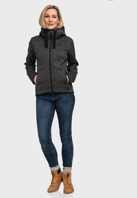 Schwarz-weiß gemusterte Jacke mit hohem Kragen, Frontreißverschluss und Taschen. Getragen mit blauen Jeans und beigefarbenen Schnürstiefeln.