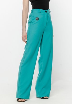 Kobieta ubrana w turkusowe spodnie o szerokich nogawkach i czarny bezrękawnik, stojąca z jedną ręką swobodnie opuszczoną wzdłuż ciała.