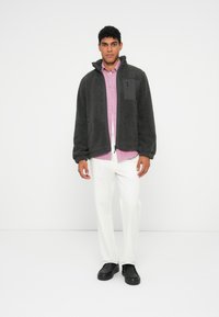 Vīrietis stāv melnā svarccio jaku virs rozā pogājamas krekla, baltām biksēm un melnām kurpēm uz balta fona.