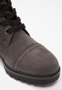 Gros plan sur une botte en daim gris foncé avec lacets noirs, œillets métalliques, bout renforcé cousu et semelle épaisse noire à rainures.
