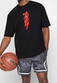 Persona vestida con una camiseta negra con texto gráfico rojo y pantalones cortos grises con estampado, sosteniendo un balón de baloncesto Spalding con una mano.