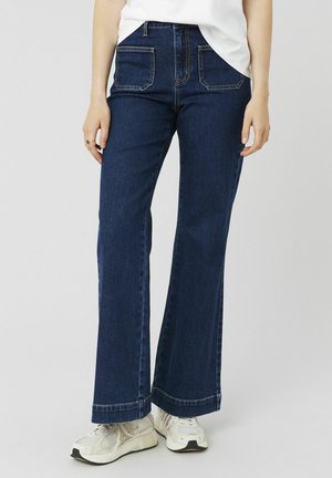 Jean flare bleu foncé en denim, avec deux poches arrière, des coutures contrastées et une taille régulière. Portés avec des baskets claires.