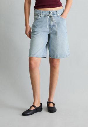 Personne portant un short en denim bleu clair jusqu'aux genoux avec une taille à cordon, un haut rouge foncé et des chaussures Mary Jane noires debout.