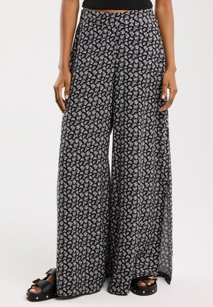 Zwarte wijde broek met een wit bloemenpatroon, gemaakt van lichtgewicht stof met een vloeiende uitstraling. Draagt met zwarte sandalen.