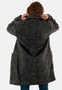 Cappotto grigio in lana con un motivo testurizzato, vestibilità oversize, ampie tasche laterali e un colletto prominente. La vista posteriore mostra i dettagli delle cuciture.