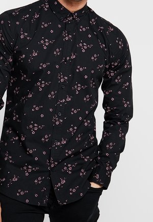 Homme portant une chemise noire à boutons avec un petit imprimé floral rose, une main dans la poche, sur un fond gris clair uni.