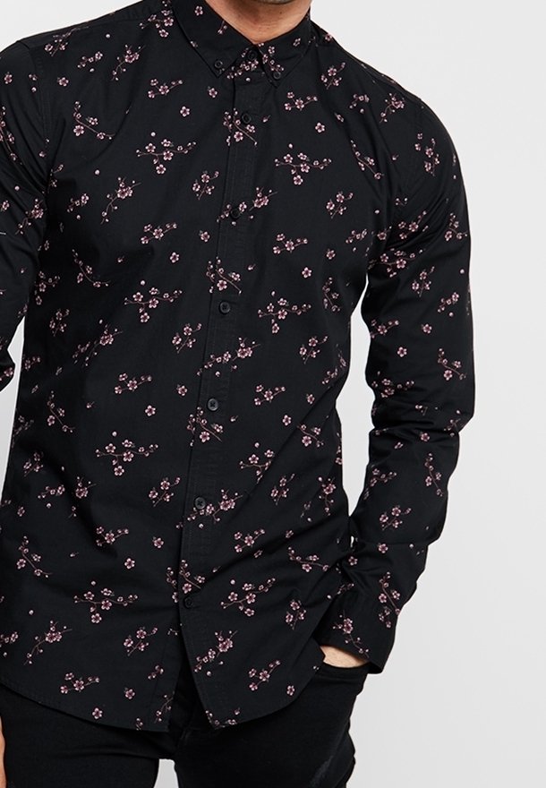 Homme portant une chemise noire à boutons avec un petit imprimé floral rose, une main dans la poche, sur un fond gris clair uni.