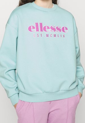Osoba nosząca jasnoniebieską bluzę z różowym napisem "ellesse EST. MCMLIX" i jasnofioletowe spodnie, ręce w kieszeniach.