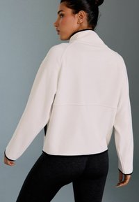Weiße Fleecejacke mit einem hohen Kragen, schwarzem Besatz an den Rändern und einer lockeren Passform, kombiniert mit schwarzen Leggings. Gleiche Textur und gemütliches Design.