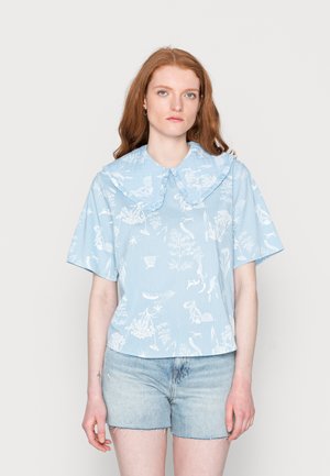 Camisa de manga curta azul riscada com um grande colarinho franzido. Apresenta motivos botânicos e animais brancos, fabricada em tecido leve. Usada com calções de ganga.
