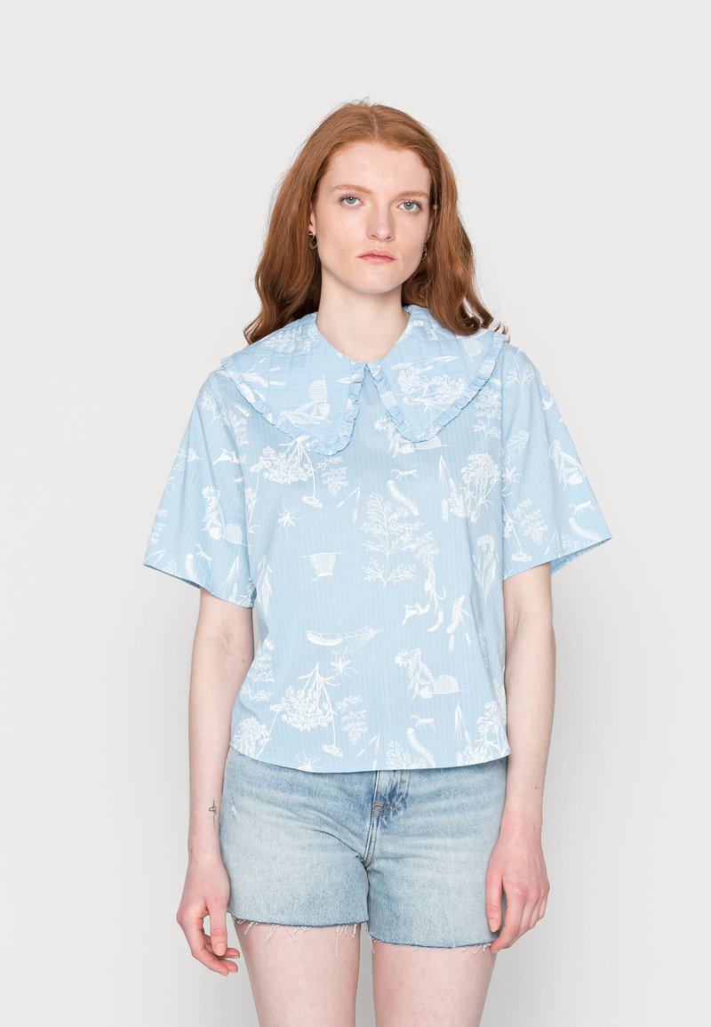 Camisa de manga curta azul riscada com um grande colarinho franzido. Apresenta motivos botânicos e animais brancos, fabricada em tecido leve. Usada com calções de ganga.