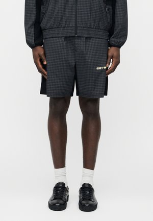 Inimene kannab musta ruudulise mustriga lühikesi pükse "ea7" logoga, sobivat jopet, valgeid sokke ja musti tossusid, seistes valgel taustal.