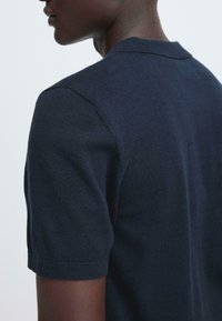 Polo bleu marine à manches courtes en tissu lisse. Présente un col et des poignets côtelés avec un détail de couture minimal.