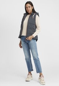 Un gilet gris rembourré avec une capuche et une fermeture éclair à l'avant, doté de boutons beiges et de poches, porté sur un pull léger et un jean bleu avec des baskets.