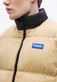 Pruun puffer-jope musta kraega, esikinnitusklapi ja sinise "HUGO" logoga sildiga. Omad quiltitud tekstuuri ja kontrastseid musti aktsente.