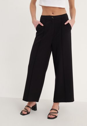 Femme portant un pantalon noir taille haute à jambes larges avec poches latérales et des sandales à talons noires à brides, mains dans les poches, top blanc court.