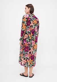 Robe maxi florale en tissu foncé ornée de fleurs roses vives, oranges et vertes. Manches longues avec poignets boutonnés et fentes latérales.