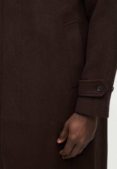 Nahaufnahme eines dunkelbraunen Wollmantelärmels mit geknöpftem Riemendetail und einer dunkelhäutigen Hand, die neben der Manteltasche ruht.