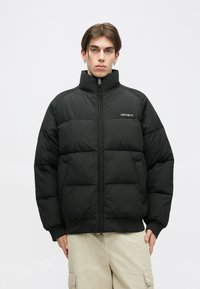 Fekete puffertöltött dzseki magas gallérral, steppelt mintázattal, elől cipzárral és logóval díszítve. Az anyaga simának és szigeteltnek tűnik, a mandzsetták pedig szoros fazonúak.