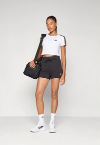 Kvinne i hvit crop top og svarte shorts med Adidas-logo, bærende en svart treningsbag, iført hvite sokker og sneakers, som står mot en ensfarget bakgrunn.