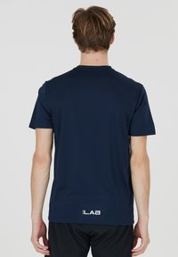 Tummansininen urheilupaita, lyhythihainen, pyöreä kaula-aukko, valmistettu sileästä kankaasta, jossa on logo helman kohdalla, minimalistinen muotoilu.